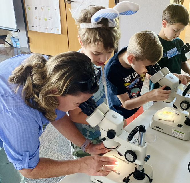 An educator uses a microscope as a child looks on, and two other children use microscopes in the background